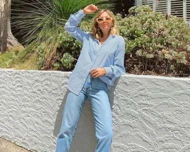 Woman in sunglasses and blue striped shirt, leaning against a white wall with plants.