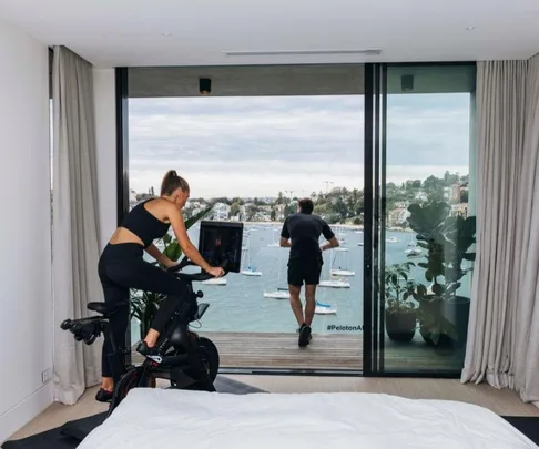 A woman exercises on a Peloton bike in a modern room with a view of a bay and boats through large glass doors.