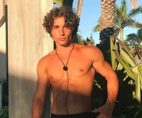 Shirtless young man with curly hair stands outdoors near tropical plants.