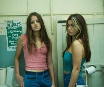 Two young women in a bathroom, one in a pink top and the other in teal, with a poster in the background.