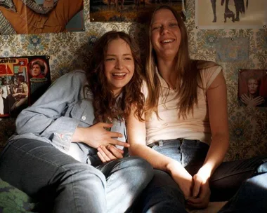 Two young women sitting and laughing on a bed, surrounded by posters, in a warmly lit room.