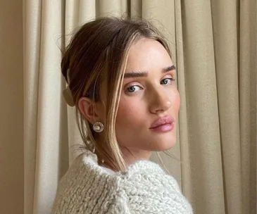 Person with light makeup and sweater, posing in front of beige curtains.