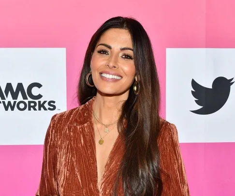 Smiling woman with long dark hair in a rust-colored top, standing in front of AMC and Twitter logos on a pink background.