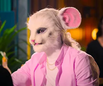 Person in a detailed mouse-like mask and pink shirt from Netflix's "Sexy Beasts," sitting indoors.