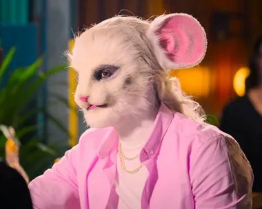 Person in a detailed mouse-like mask and pink shirt from Netflix's "Sexy Beasts," sitting indoors.