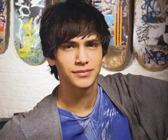 Young man with brown hair in a blue shirt and gray jacket stands in front of graffiti-covered skateboards.