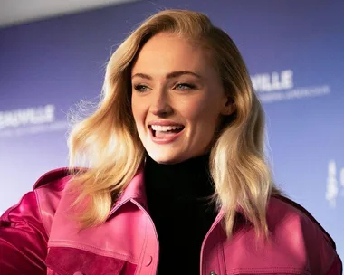 A person smiling, wearing a pink jacket, with long blonde hair against a blurred blue background.