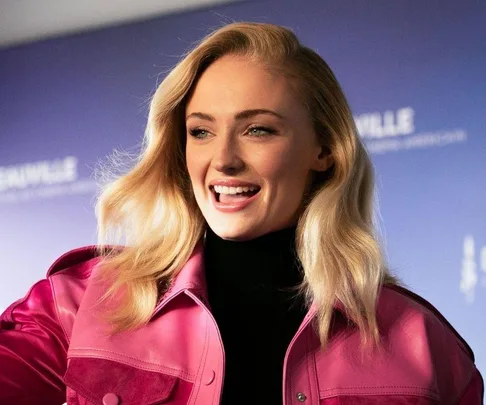 A person smiling, wearing a pink jacket, with long blonde hair against a blurred blue background.