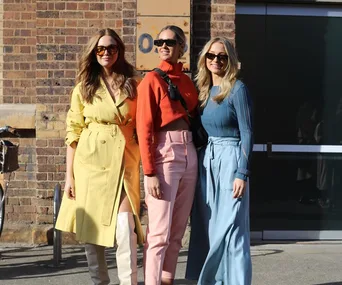 Three women in bright outfits at Australian Fashion Week 2021; one in yellow, one in orange and pink, one in blue.