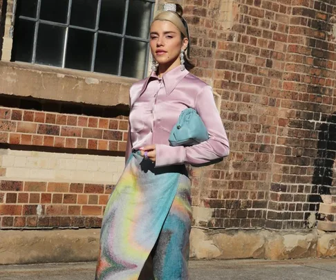 Woman in a pink satin blouse and colorful skirt, holding a blue purse, standing against a brick wall.