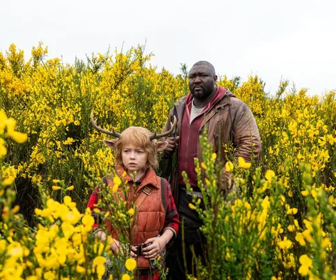 A boy with antlers and a man stand in a field of yellow flowers from the series "Sweet Tooth."