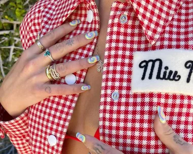 Hands with tie-dye nails on a red checkered Miu Miu jacket, adorned with rings.