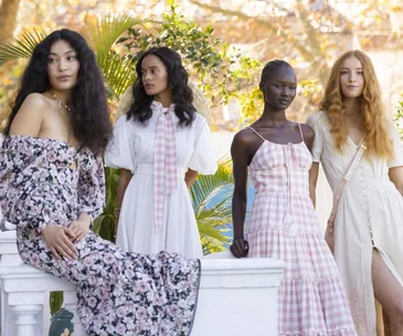 Four women in floral and gingham dresses pose together outdoors by a white railing with lush greenery in the background.