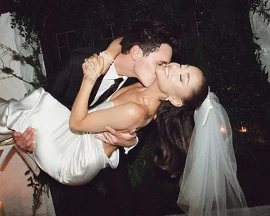 Bride in white gown and veil being affectionately lifted by groom in dark suit during their wedding celebration.