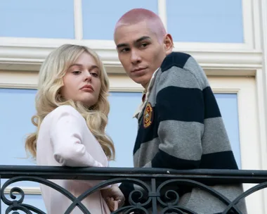 Two characters from Gossip Girl reboot standing on a balcony, one with long blonde hair, the other with short pink hair.