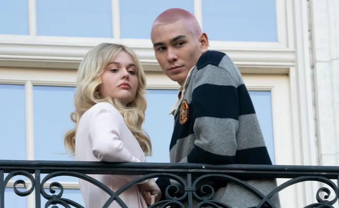 Two characters from Gossip Girl reboot standing on a balcony, one with long blonde hair, the other with short pink hair.