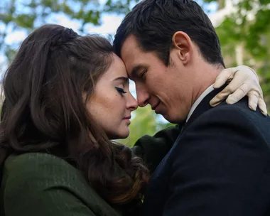 A couple standing close with their foreheads touching in a tender moment, outdoors with blurred green foliage in the background.