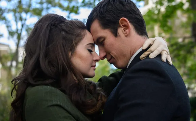 A couple standing close with their foreheads touching in a tender moment, outdoors with blurred green foliage in the background.