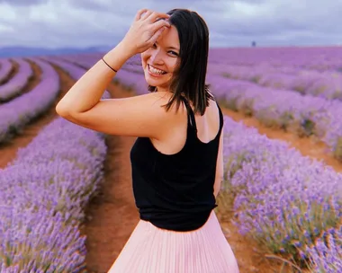 A woman smiling in a lavender field, wearing a black tank top and pink skirt.