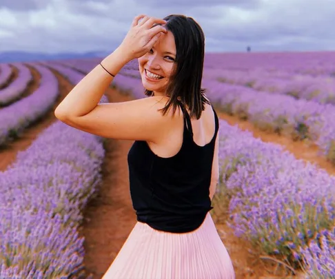 A woman smiling in a lavender field, wearing a black tank top and pink skirt.