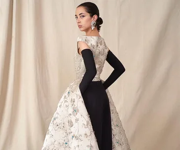 Elegant woman in embroidered gown with black gloves, looking back over her shoulder, standing against a neutral backdrop.