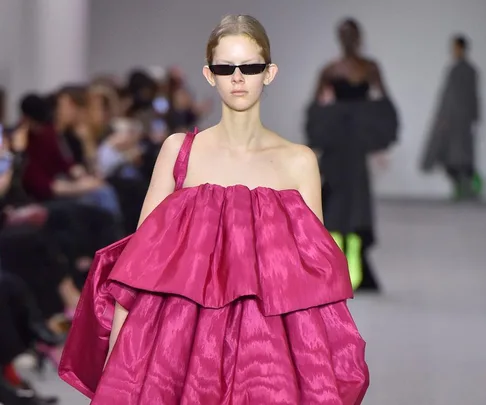 Model in a strapless, voluminous pink dress and sunglasses walks a runway with blurred audience background.