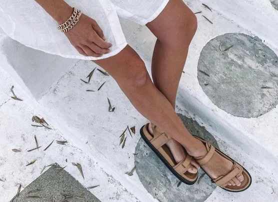 Woman wearing tan leather sandals and white dress, sitting on stone steps outdoors.