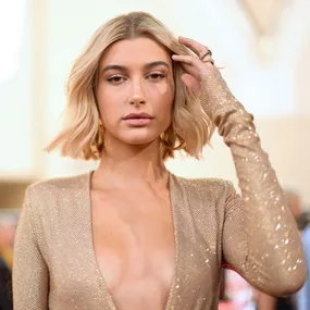 A woman in a sheer, sparkling gold dress touches her short, blonde hair, posing confidently at an event.