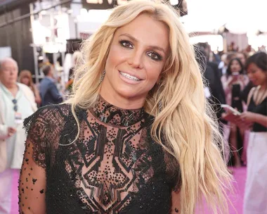 Woman in a black lace dress smiles at a public event with a crowd in the background.