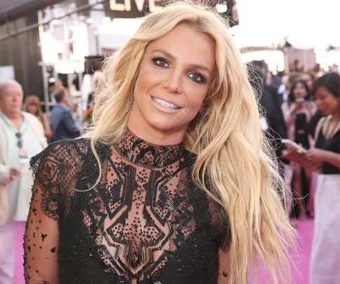 Woman in a black lace dress smiles at a public event with a crowd in the background.