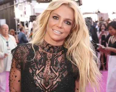 Blonde woman in a black lace dress smiles at a glamorous event with people and cameras in the background.