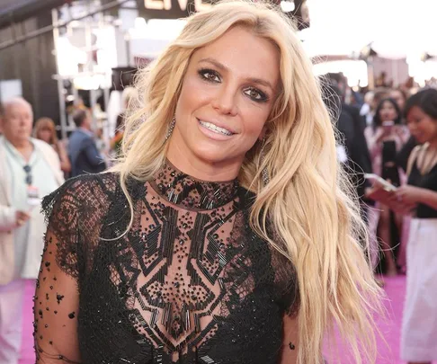 Blonde woman in a black lace dress smiles at a glamorous event with people and cameras in the background.