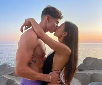 A couple kisses passionately near a body of water during sunset, capturing a romantic moment.