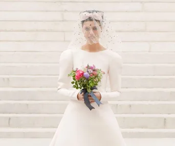 Model in a white wedding dress and veil holds a colorful bouquet, standing on stone steps at Chanel Fall/Winter 2021.