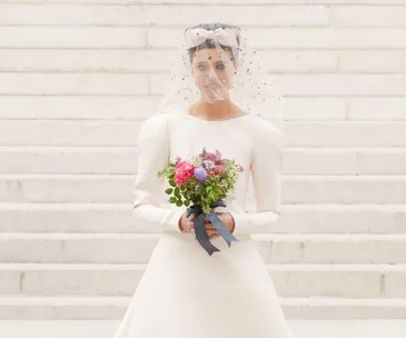 Model in a white wedding dress and veil holds a colorful bouquet, standing on stone steps at Chanel Fall/Winter 2021.