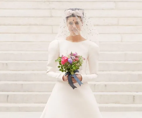 Model in a white wedding dress and veil holds a colorful bouquet, standing on stone steps at Chanel Fall/Winter 2021.