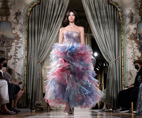 Model on runway in ornate room wearing a strapless, multicolored, voluminous gown during Couture Week Autumn/Winter 2021.