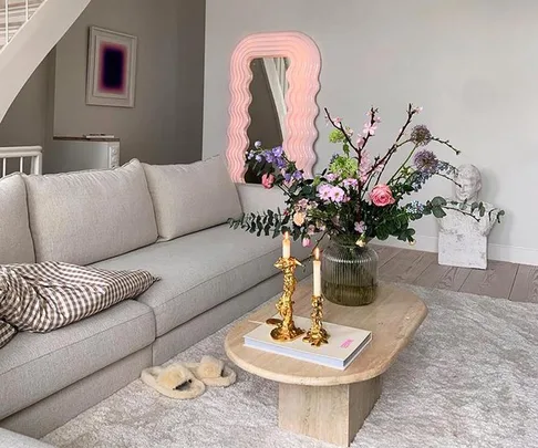 Cozy living room with a gray sofa, colorful flowers on table, gold candle holders, and a wavy pink mirror on the wall.