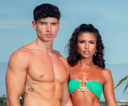 A muscular man and woman in green swimsuits stand outdoors against a clear sky background.