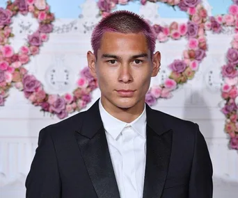 Person with pink hair in formal attire, standing before a floral backdrop with heart-shaped rose arrangements.