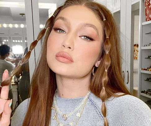 "Gigi Hadid with braided hair, wearing a grey top and layered necklaces, in a bathroom setting."