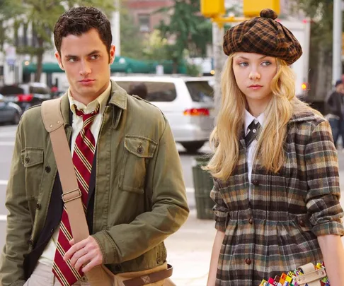 Two young people in stylish outfits walk on a city street; the woman wears a plaid dress and beret, the man a tie and jacket.