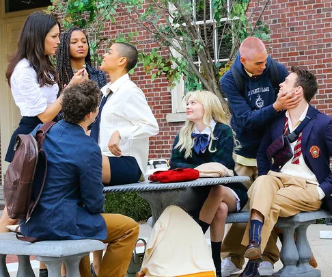 A group of young adults in school uniforms socializing on a bench outside, near a brick building and trees.