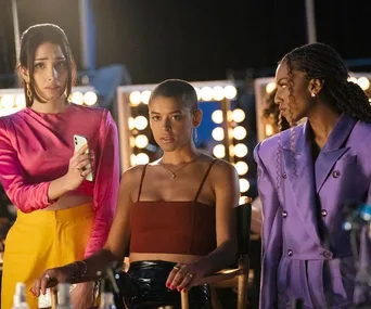 Three women on a dimly lit set, one holding a phone. They're dressed in colorful outfits, with lights in the background.