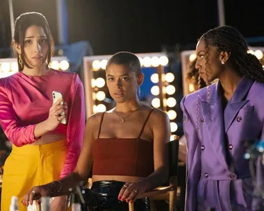 Three women on a dimly lit set, one holding a phone. They're dressed in colorful outfits, with lights in the background.