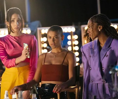 Three women on a dimly lit set, one holding a phone. They're dressed in colorful outfits, with lights in the background.