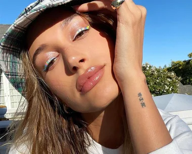 Woman with glossy lips, wearing plaid hat and rainbow eyeliner stickers, poses outdoors.