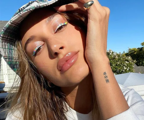 Woman with glossy lips, wearing plaid hat and rainbow eyeliner stickers, poses outdoors.