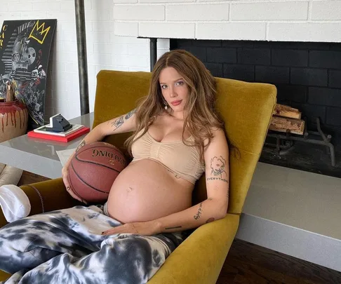 Pregnant woman in lounge chair holding a basketball, with tattoos visible, near a fireplace and art pieces.