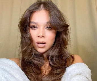 Woman with long, wavy brown hair and neutral makeup, wearing an off-shoulder white garment, in front of a beige background.